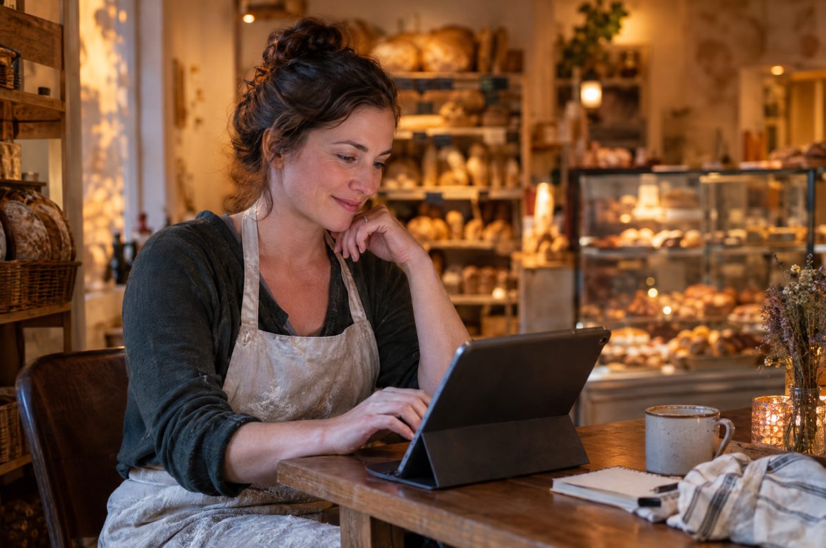 A bakery owner in an apron smiles as she types on a tablet at a corner table of her shop after closing, with bread shelves and pastry cases behind her.